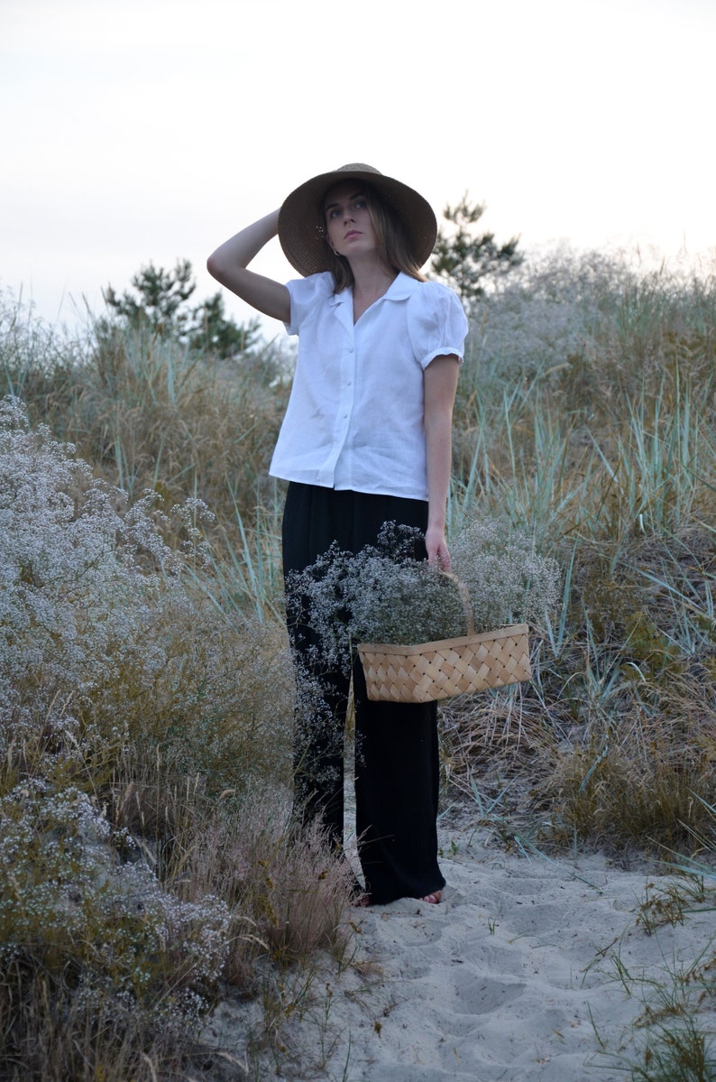 May include: A woman wearing a white linen shirt and black pants holds a woven basket filled with white flowers. She is standing in a field of tall grass and wildflowers.