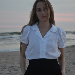 May include: A woman wearing a white collared shirt with puffed sleeves and a black skirt, standing on a sandy beach with the ocean in the background.
