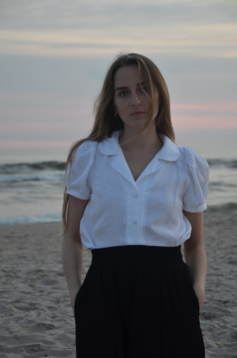 May include: A woman wearing a white collared shirt with puffed sleeves and a black skirt, standing on a sandy beach with the ocean in the background.