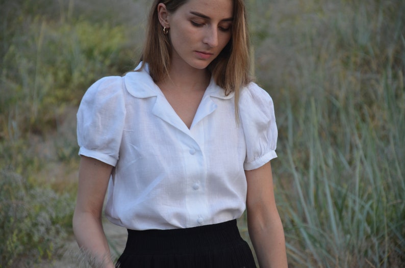 May include: A white, collared blouse with puffed sleeves and a button-up front. The blouse is made of a lightweight fabric and has a relaxed fit.