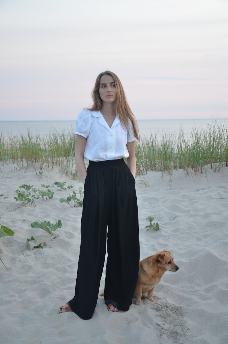 May include: A woman wearing a white collared shirt and black wide-leg pants stands on a sandy beach with a small brown dog.