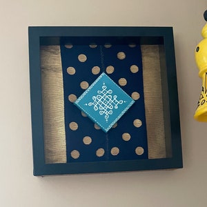 May include: Framed artwork with a teal and white geometric design on a navy blue polka dot background. The artwork is set against a gold fabric backdrop within a black frame. A yellow decorative bell hangs nearby.