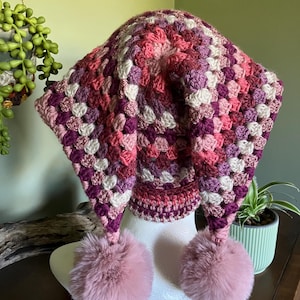 May include: A colourful crochet hat in pinks, purples, and white, with a granny square pattern. The hat has long ear flaps and is decorated with two fluffy pink pom-poms. This unique hat is a cosy and stylish accessory.