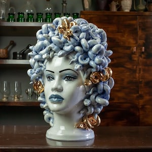May include: A white ceramic sculpture of a woman's head with a blue and gold snake hairdo. The sculpture is on a wooden surface.