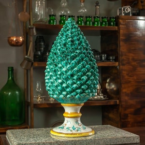 Puede incluir: Una escultura de cerámica en forma de piña con un esmalte verde y una base blanca y amarilla. La base está decorada con diseños florales.