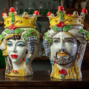 Puede incluir: Dos jarrones de cerámica con forma de cabeza con coloridos diseños florales y de frutas. Los jarrones están decorados con coronas y tienen un fondo blanco con detalles amarillos, verdes, rojos y azules.