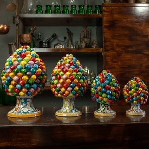 Op de afbeelding: Vier kleurrijke keramische dennenappelsculpturen in verschillende maten. De dennenappels zijn versierd met verschillende kleuren, waaronder rood, geel, groen en blauw. De sculpturen zijn in een rij op een houten tafel geplaatst.