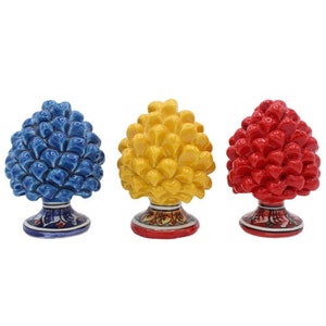 Può includere: Tre statuette in ceramica a forma di pigna, blu, gialle e rosse. Ognuna ha una superficie dettagliata e strutturata e poggia su una base decorativa con motivi floreali dipinti. Su sfondo bianco.