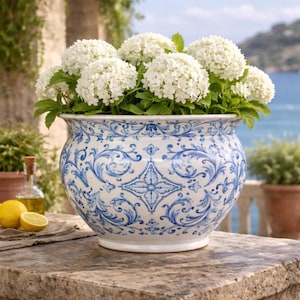 Op de afbeelding: Een grote, ronde keramische plantenbak met een blauw en wit bloemmotief, gevuld met witte hortensia's. De plantenbak staat op een stenen oppervlak, met een kustachtergrond zichtbaar.