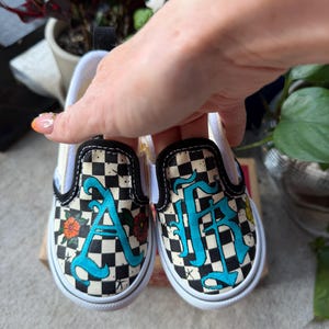 Könnte beinhalten: Ein Paar Slipper mit schwarz-weißem Schachbrettmuster. Die Schuhe sind mit handgemalten, türkisfarbenen Buchstaben im gotischen Stil verziert, die durch kleine orangefarbene und gelbe Blumenmotive sowie schwarze Sternendetails ergänzt werden.