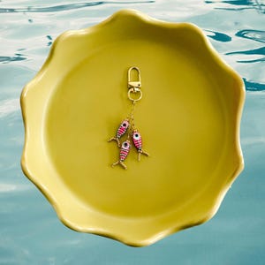 May include: A gold-toned metal keychain featuring three small, dangling fish charms. Each fish is crafted from tiny beads in shades of pink, white, and black, with a prominent eye detail. The charms are attached to a gold-toned lobster clasp.