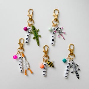 May include: Five gold-toned metal keychains featuring beaded animal charms and white letter beads spelling LATER, MOODY, SILLY, BUSY, and FERAL. Each keychain includes a colorful round accent bead and a lobster clasp for attachment to bags or keys.
