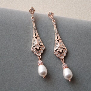 May include: A pair of rose gold drop earrings. Each earring has an ornate, openwork design with clear crystals and a pear-shaped white pearl. The earrings have a vintage, elegant style.