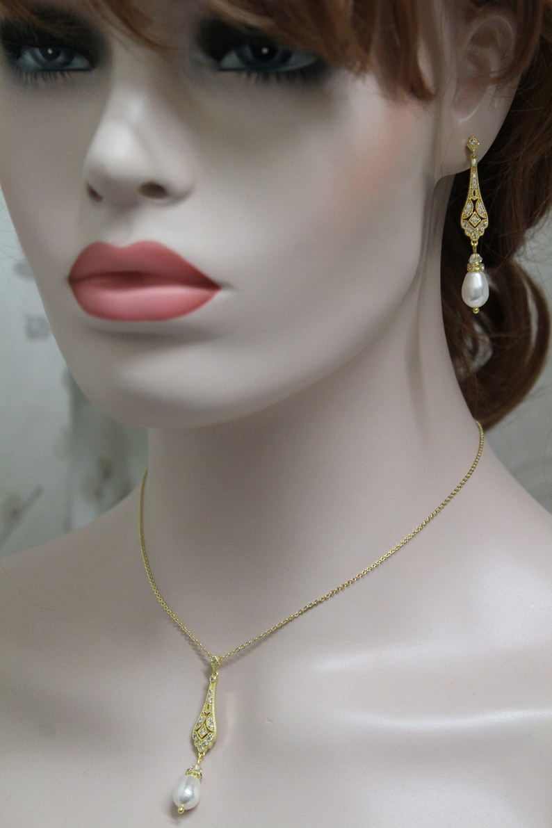 May include: A gold necklace and earrings set with a teardrop-shaped pendant and matching earrings. The pendant and earrings are adorned with clear crystals and a single pearl drop.