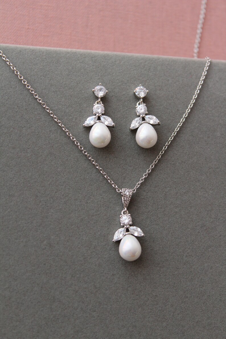 May include: A silver necklace and earrings set with a pearl and crystal accents. The necklace has a delicate chain and a pendant with a pearl drop and crystal accents. The earrings have a pearl drop and crystal accents.
