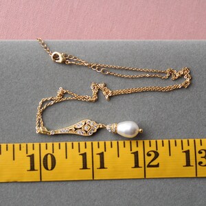 May include: A gold chain necklace with a delicate pendant featuring a pearlescent drop and a geometric design with clear stones. The necklace is measured against a yellow tape measure.