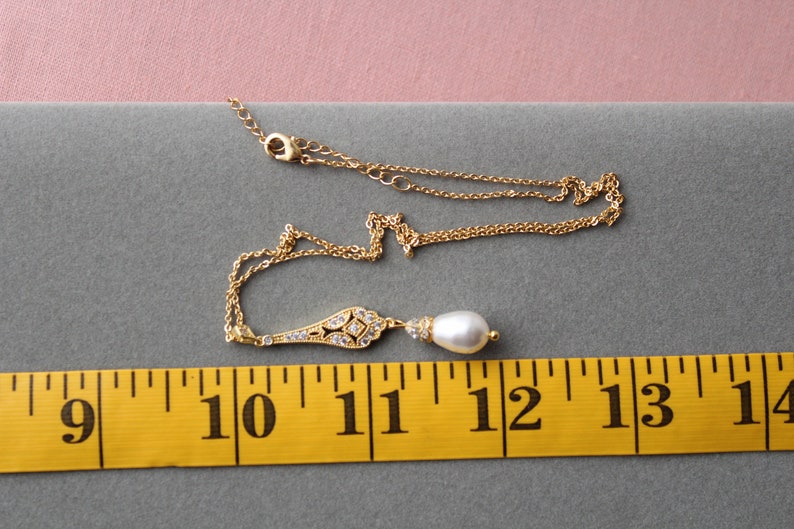 May include: A gold chain necklace with a delicate pendant featuring a pearlescent drop and a geometric design with clear stones. The necklace is measured against a yellow tape measure.