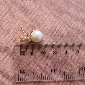 Puede incluir: Pendientes de tono dorado con una perla y detalles de cristal transparente. Los pendientes se miden con una regla, mostrando que la perla tiene aproximadamente 1 cm de di&aacute;metro.