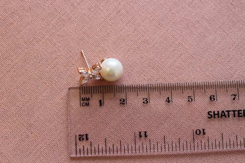 Puede incluir: Pendientes de tono dorado con una perla y detalles de cristal transparente. Los pendientes se miden con una regla, mostrando que la perla tiene aproximadamente 1 cm de di&aacute;metro.