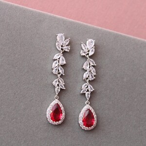 May include: A pair of silver dangle earrings featuring a red teardrop-shaped stone at the bottom. The earrings are decorated with clear cubic zirconia stones in a leaf-like pattern.