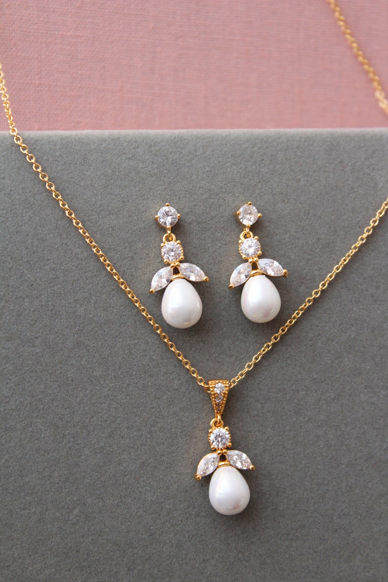 May include: A gold necklace and earrings set with a pearl pendant and matching pearl drop earrings. The pendant and earrings feature a cluster of clear cubic zirconia stones.