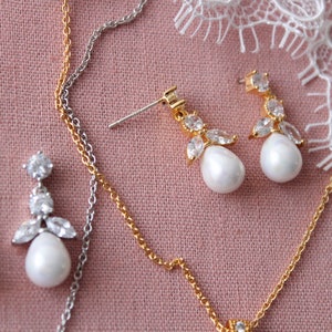 May include: Two pairs of pearl and crystal earrings on a pink fabric background. One pair has a silver chain and the other has a gold chain.