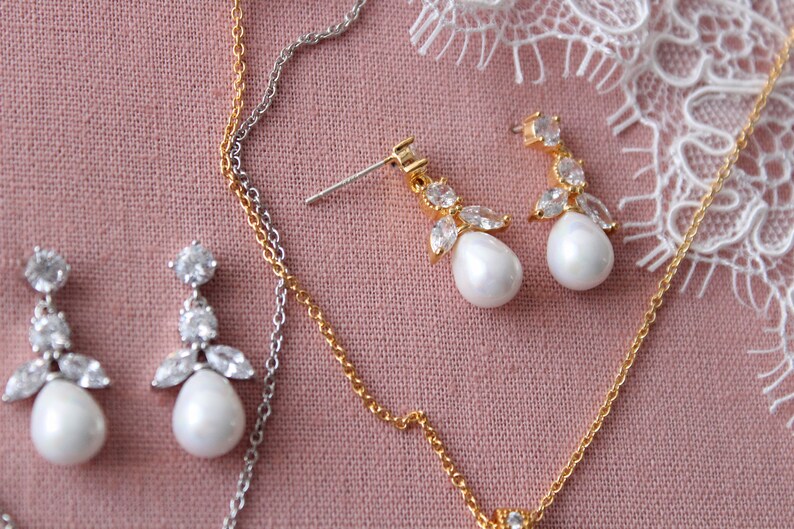 May include: Two pairs of pearl and crystal earrings on a pink fabric background. One pair has a silver chain and the other has a gold chain.