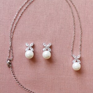 Puede incluir: Un collar de plata con un colgante de perla y pendientes de perla a juego. El colgante y los pendientes presentan un peque&ntilde;o dise&ntilde;o de flor de cristal transparente.