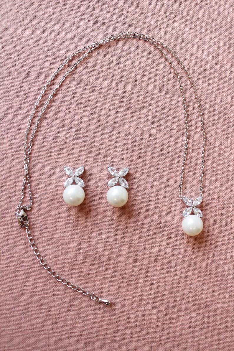 Puede incluir: Un collar de plata con un colgante de perla y pendientes de perla a juego. El colgante y los pendientes presentan un peque&ntilde;o dise&ntilde;o de flor de cristal transparente.