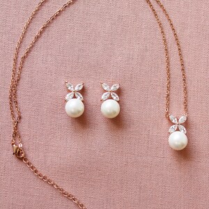 Puede incluir: Un collar y pendientes de oro rosa con un colgante de perla y pendientes de perla a juego. El colgante y los pendientes presentan un delicado dise&ntilde;o floral con piedras de circonita c&uacute;bica transparentes.