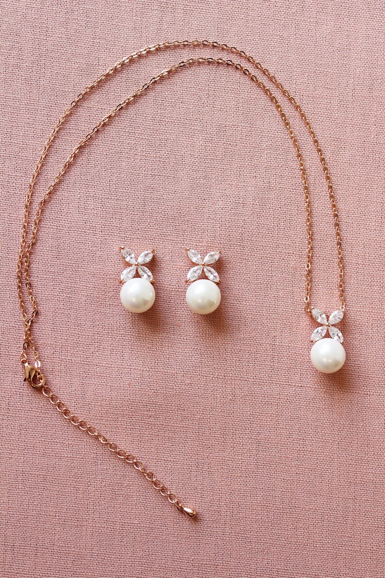 Puede incluir: Un collar y pendientes de oro rosa con un colgante de perla y pendientes de perla a juego. El colgante y los pendientes presentan un delicado dise&ntilde;o floral con piedras de circonita c&uacute;bica transparentes.