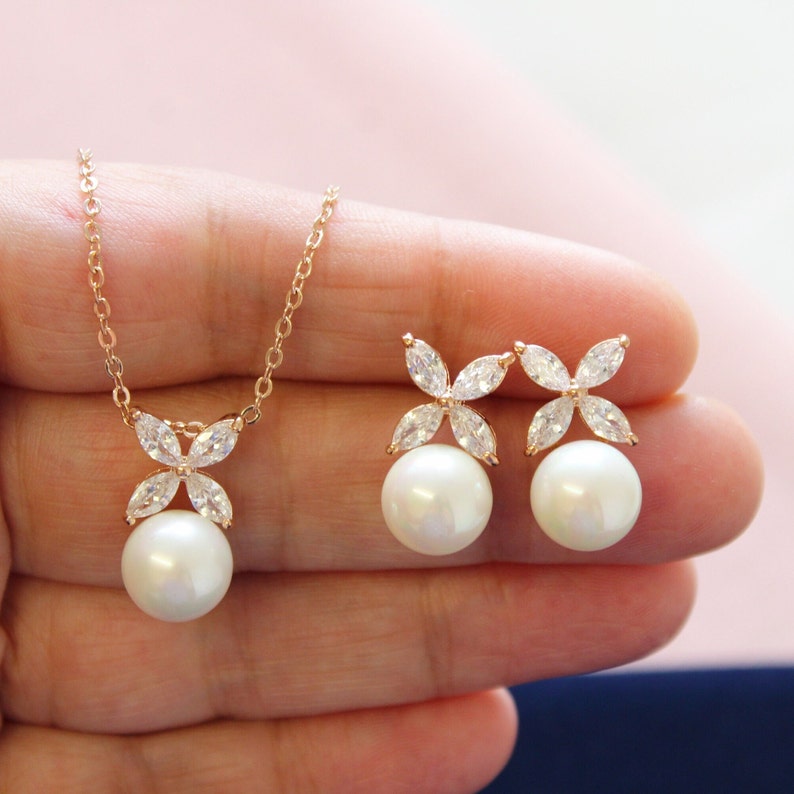 Puede incluir: Un conjunto de collar y pendientes de oro con un colgante de flor de cristal y perla. El collar tiene una cadena delicada y los pendientes son de estilo de bot&oacute;n.