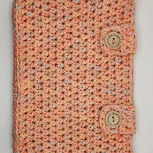 May include: A crocheted peach-colored book cover with a light yellow and gray speckled pattern. The cover has two wooden buttons with the text "Handmade" on them.