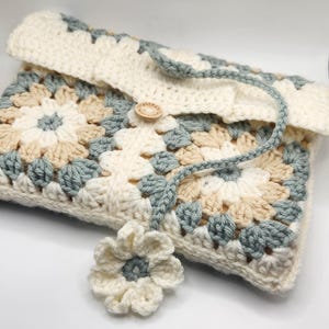 Puede incluir: Una bolsa de ganchillo con un diseño floral en tonos de blanco, beige y azul. La bolsa tiene un botón de madera con la palabra "Handmade" y una flor de ganchillo unida a la correa.