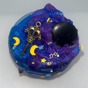 May include: A jar of blue and purple slime with gold, yellow, and blue embellishments. The slime is decorated with a gold key, yellow crescent moons, stars, and a black sphere.