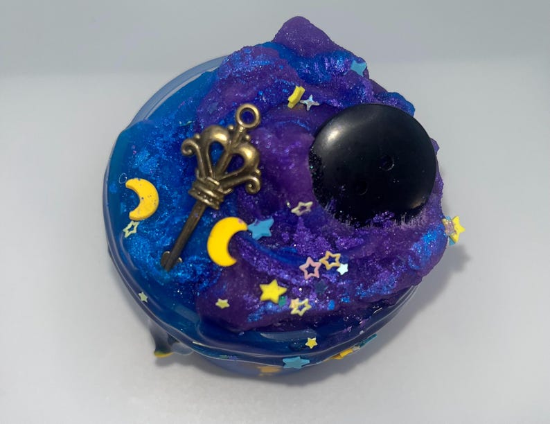 May include: A jar of blue and purple slime with gold, yellow, and blue embellishments. The slime is decorated with a gold key, yellow crescent moons, stars, and a black sphere.