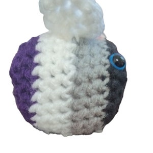 May include: A crocheted ball in a colour scheme of white, grey, black, and purple. It features a large, blue-eyed, black bead eye. A fluffy white pom-pom is attached to the top. The item is likely a handmade toy or decorative item.