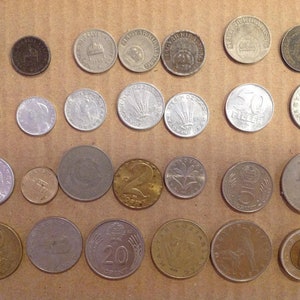 May include: A collection of 25 coins from various countries, including silver, gold, and copper coins. The coins are arranged in rows on a brown surface.