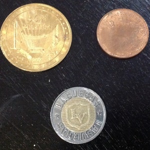 May include: Three different coins, one gold, one silver, and one copper. The gold coin has a design of a rocket ship and the words "50th Anniversary" on it. The silver coin has the words "Las Vegas" and "The Strip" on it. The copper coin has the words "Souvenir Token" on it.