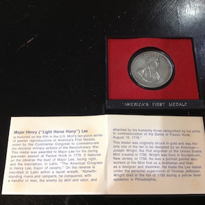 May include: A silver medal displayed in a red velvet-lined case. The medal shows a man's bust and the text "America's First Medals." The accompanying text provides details about the medal's history and Major Henry Lee.