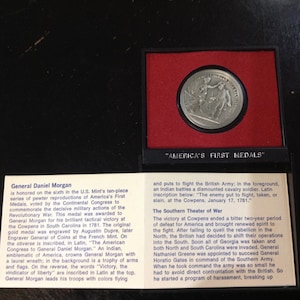 May include: A silver medal commemorating General Daniel Morgan, a hero of the American Revolutionary War. The medal depicts General Morgan leading troops, with an inscription that reads "The enemy put to flight, taken, or slain, at the Cowpens, January 17, 1781."