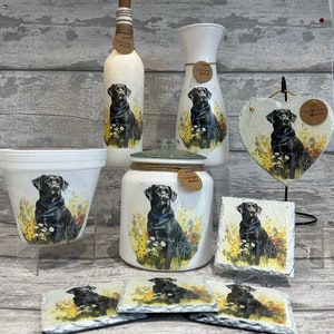 May include: Set of black Labrador dog themed home decor items. Includes a vase, a bottle, a jar, a planter, a heart-shaped slate, and four coasters. All items feature a black Labrador dog sitting in a field of flowers. Each item has a brown tag with the text "Handmade with love".
