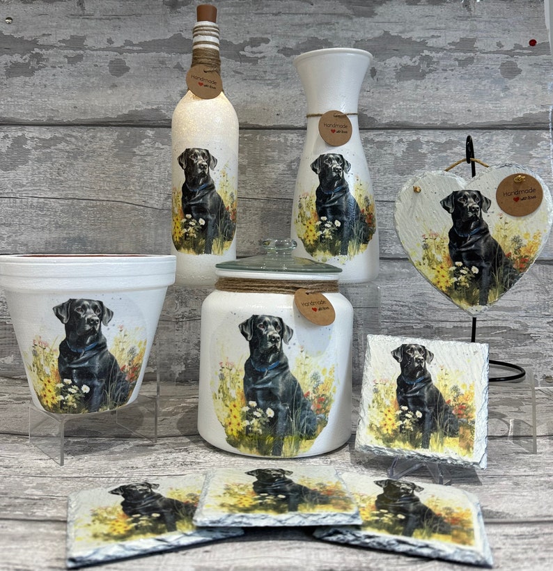 May include: Set of black Labrador dog themed home decor items. Includes a vase, a bottle, a jar, a planter, a heart-shaped slate, and four coasters. All items feature a black Labrador dog sitting in a field of flowers. Each item has a brown tag with the text "Handmade with love".