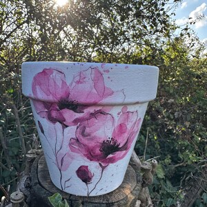 Könnte beinhalten: Weißer Keramik-Blumentopf mit handgemaltem Design von rosa Mohnblumen. Der Topf ist zylindrisch mit leicht ausgestelltem Rand und steht auf einem Baumstumpf. Das Mohnblumen-Design umfasst detaillierte Blütenblätter und Stiele.