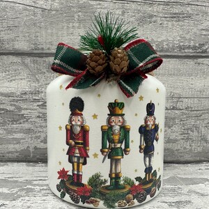May include: White ceramic jar decorated with a green and red tartan ribbon and a sprig of green foliage with pine cones. The jar features a Christmas scene with three nutcracker figures, one in red, one in green, and one in blue.
