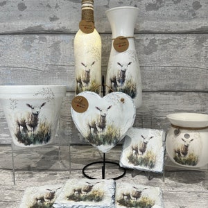 May include: A set of white ceramic home decor items featuring a hand-painted design of a lamb in a field. The set includes a vase, a bottle, a planter, a heart-shaped ornament, a candle holder, and four coasters. Each item has a brown tag that says "Handmade with love".