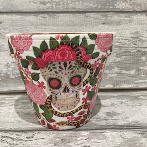 Könnte beinhalten: Weißer Keramik-Blumentopf mit einem farbenfrohen Zucker Schädel Design und einem Schlangenmuster. Der Topf ist mit rosa Blumen und grünen Blättern verziert.