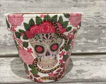 Peut inclure: Pot de fleurs en céramique blanche avec un motif de crâne de sucre coloré et un motif de serpent. Le pot est décoré de fleurs roses et de feuilles vertes.