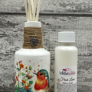 May include: White diffuser bottle with a floral and bird design, a brown twine bow, and a silver heart charm. The bottle is filled with wooden diffuser sticks. A white bottle of "Fresh Linen Diffuser Refill" by "ickletickle home scents" is next to the diffuser bottle.