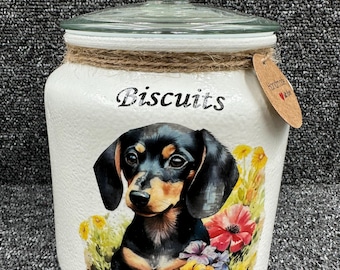 May include: White ceramic cookie jar with a glass lid. The jar features a hand-painted image of a dachshund puppy sitting in a field of flowers. The jar is labeled "Biscuits".
